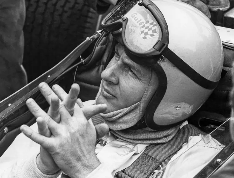 Black-and-white photo of Bruce McLaren in his helmet, seated in the cockpit