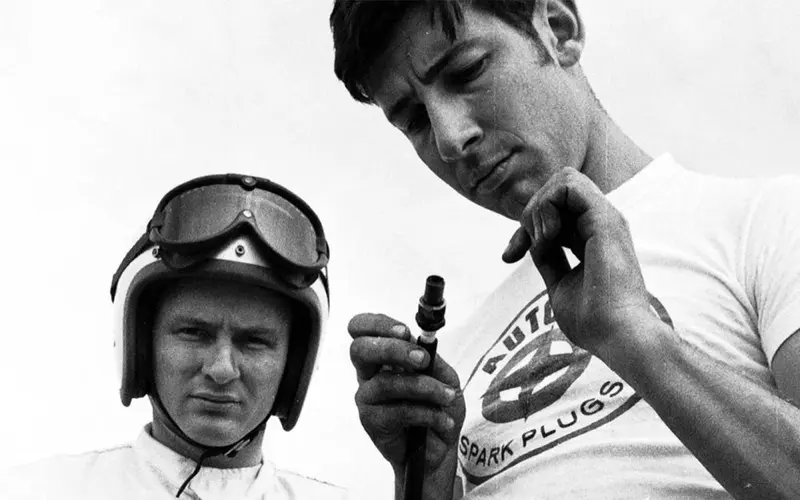 Black-and-white photo of Bruce McLaren in his helmet beside a team member inspecting a spark plug