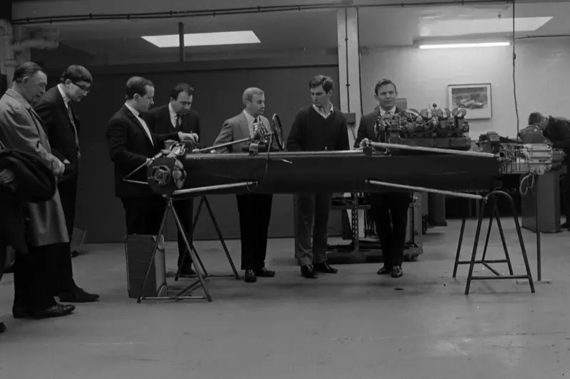 Black-and-white photo of the early McLaren team gathered around an engine in the workshop