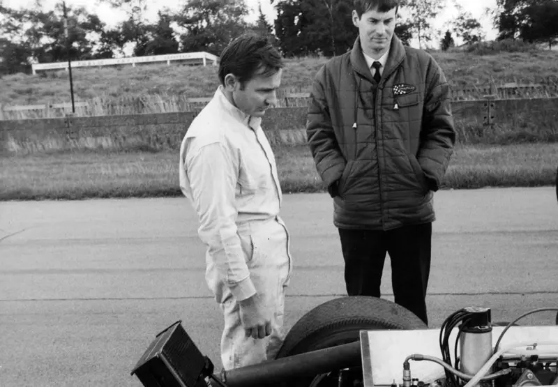 Black-and-white photo of Bruce McLaren and a colleague studying a car at the circuit