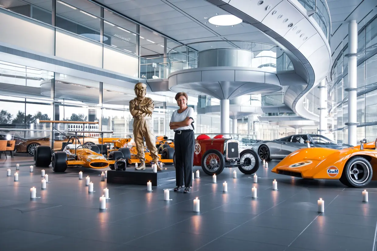 Amanda McLaren beside a statue of her father, Bruce McLaren, surrounded by candles and historic McLaren cars at the McLaren Technology Centre