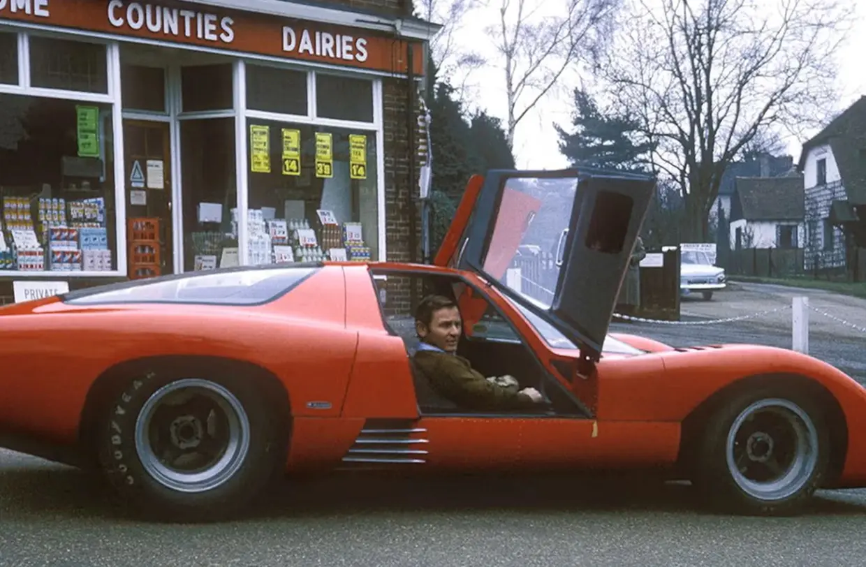 The papaya-red McLaren M6GT parked on an English high street with its door open