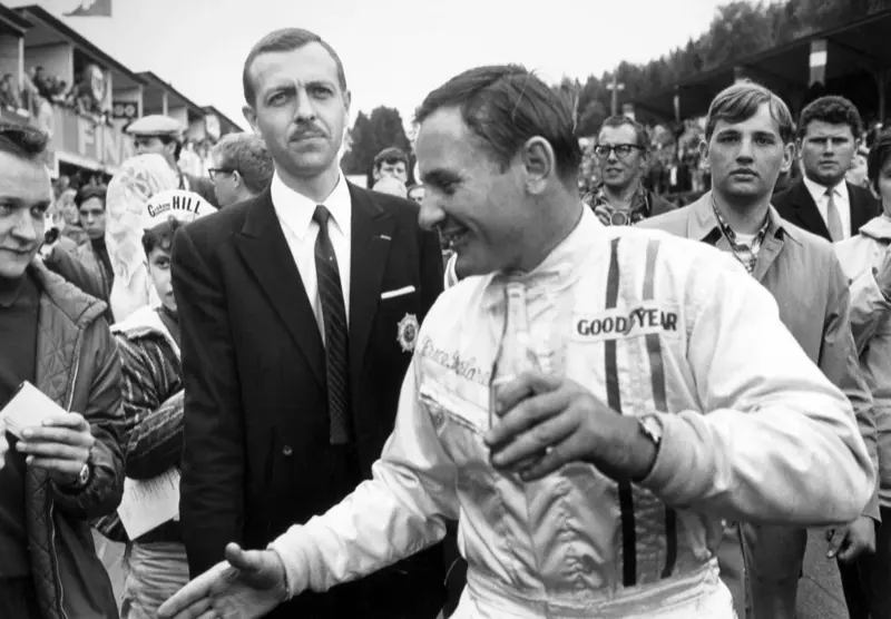 Black-and-white photo of Bruce McLaren in Goodyear overalls, smiling among a crowd