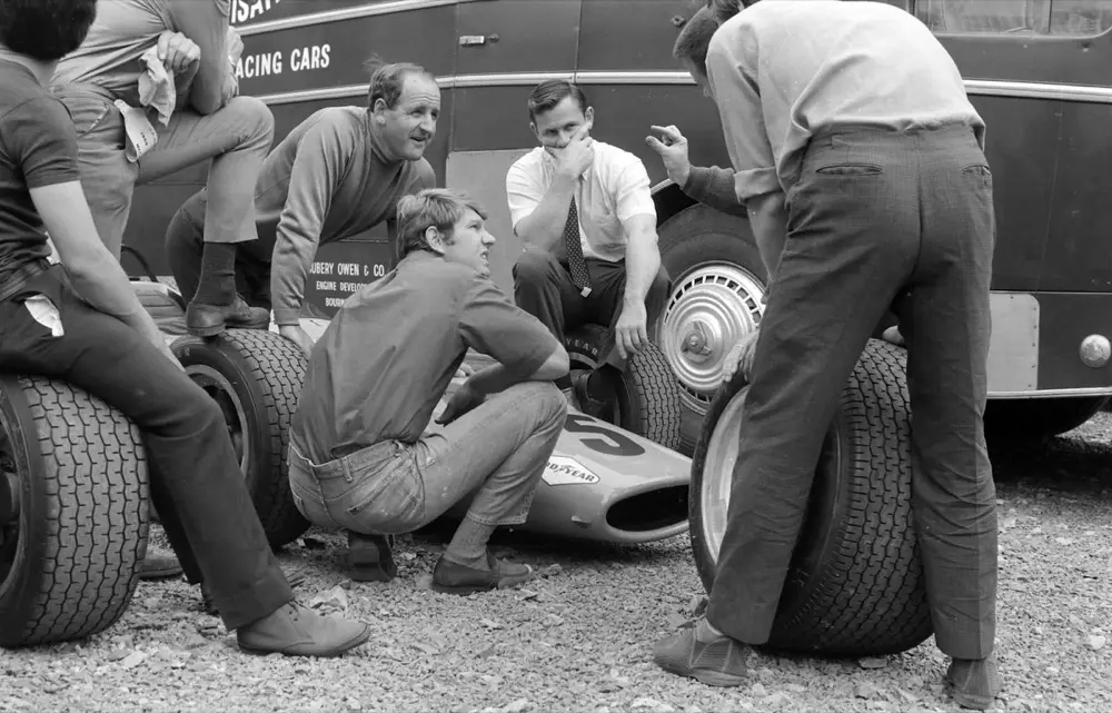 Black-and-white photo of McLaren mechanics resting on racing tyres beside the team transporter