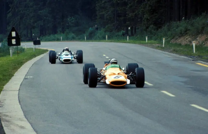 A papaya McLaren M7A leading another Formula 1 car along a tree-lined circuit